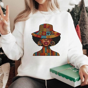 May include: White sweatshirt featuring a colorful graphic of a woman with a patterned hat, earrings, and necklace. The graphic uses a variety of geometric shapes and colors, including red, orange, yellow, and blue. The design is on the front of the sweatshirt.