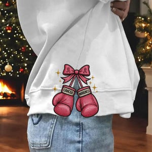 May include: White sweatshirt with a graphic of pink boxing gloves, a pink ribbon, and the words "Cancer Awareness." The design includes gold star accents. The sweatshirt is paired with blue jeans.