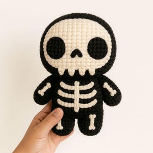 May include: A black and cream crochet skeleton doll. The doll has a skull-shaped head with large eye sockets and a jawline. The body has cream-coloured bone detailing on a black background. The doll is held in a hand.