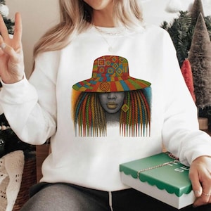 Może przedstawiać: Biała bluza z kolorową grafiką kobiety z długimi, zaplecionymi włosami i wzorzystym kapeluszem. Grafika zawiera żywe odcienie czerwieni, pomarańczy, żółci, zieleni i niebieskiego. Twarz kobiety jest w odcieniach złota, z pasującymi kolczykami.