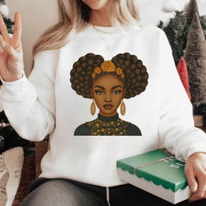 Op de afbeelding: Witte sweatshirt met een afbeelding van een zwarte vrouw met bruine afro-plukken, gouden oorbellen en een bloemenhoofddeksel. De top van de vrouw heeft een zwarte kraag met gouden bloemendetails. Een casual, alledaags kledingstuk.