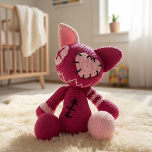 Puede incluir: Muñeca de gato de ganchillo magenta hecha a mano con un parche en forma de corazón y parches en forma de cruz. La muñeca tiene costuras blancas, puntadas negras y una pequeña bola rosa. La muñeca está sentada sobre una alfombra blanca.