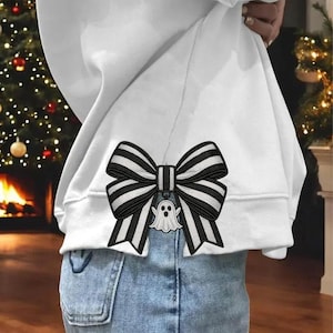 May include: A white sweatshirt with a black and white striped bow applique. The bow has a small white ghost pendant. The sweatshirt is worn with blue jeans. The design is likely Halloween-themed.