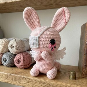 May include: A handmade pink crochet bunny with an eye patch, black button eyes, and small white wings. The bunny is sitting on a wooden shelf next to several balls of yarn in neutral colors. A thimble sits on the shelf.