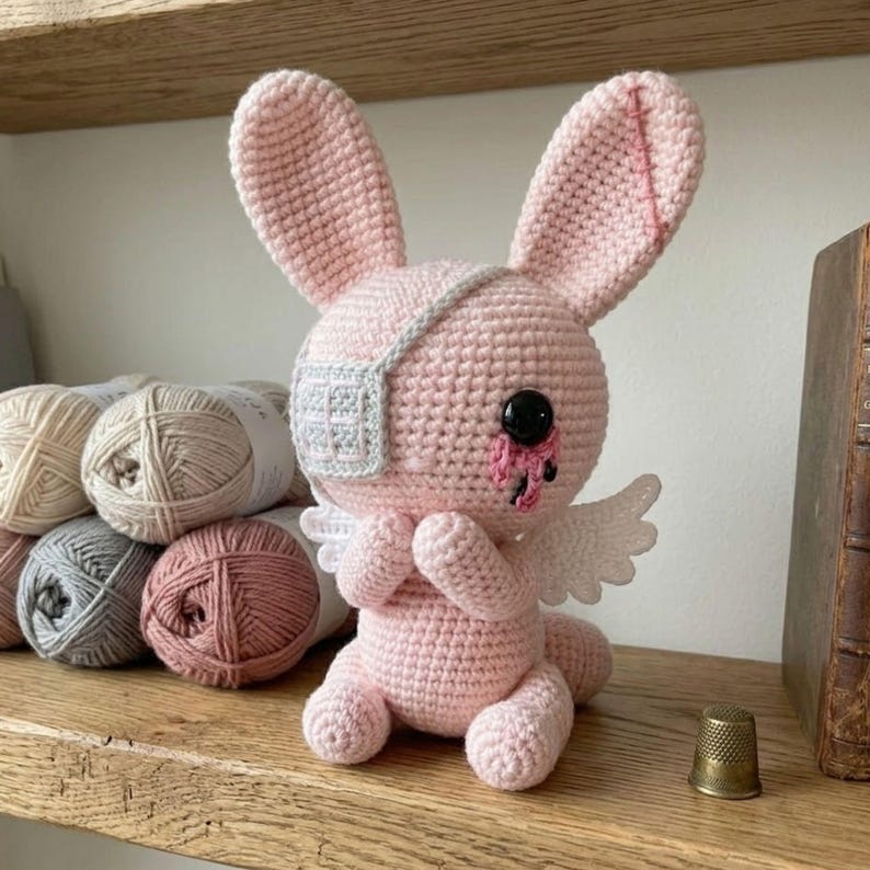 May include: A handmade pink crochet bunny with an eye patch, black button eyes, and small white wings. The bunny is sitting on a wooden shelf next to several balls of yarn in neutral colors. A thimble sits on the shelf.