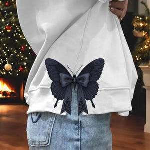 May include: A white sweatshirt with a large, dark blue butterfly design on the back. The butterfly has detailed wing patterns and a bow in the center. The sweatshirt is paired with blue jeans.