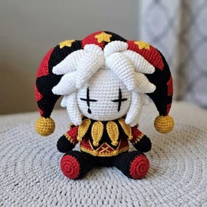 Könnte beinhalten: Gehäkelte Narrenpuppe mit weißem Gesicht, schwarzen kreuzförmigen Augen und weißen Haaren. Die Puppe trägt ein rot-schwarz-goldenes Outfit mit passendem Hut, der mit gelben Sternen und goldenen Bommeln verziert ist. Auf einer strukturierten, hellgrauen Oberfläche.