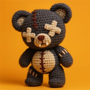 Könnte beinhalten: Ein handgehäkelter Teddybär in Dunkelgrau mit beigen Akzenten. Der Bär hat kreuzförmige Flicken über den Augen und ein gesticktes Design auf Kopf und Bauch. Nase und Mund des Bären sind braun, mit kleinen weißen Zähnen.