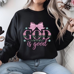 Può includere: Felpa nera con la scritta "GOD is good" in rosa e design floreale. Il design include un fiocco rosa e piccoli accenti di stelle blu. La felpa è uno stile girocollo.