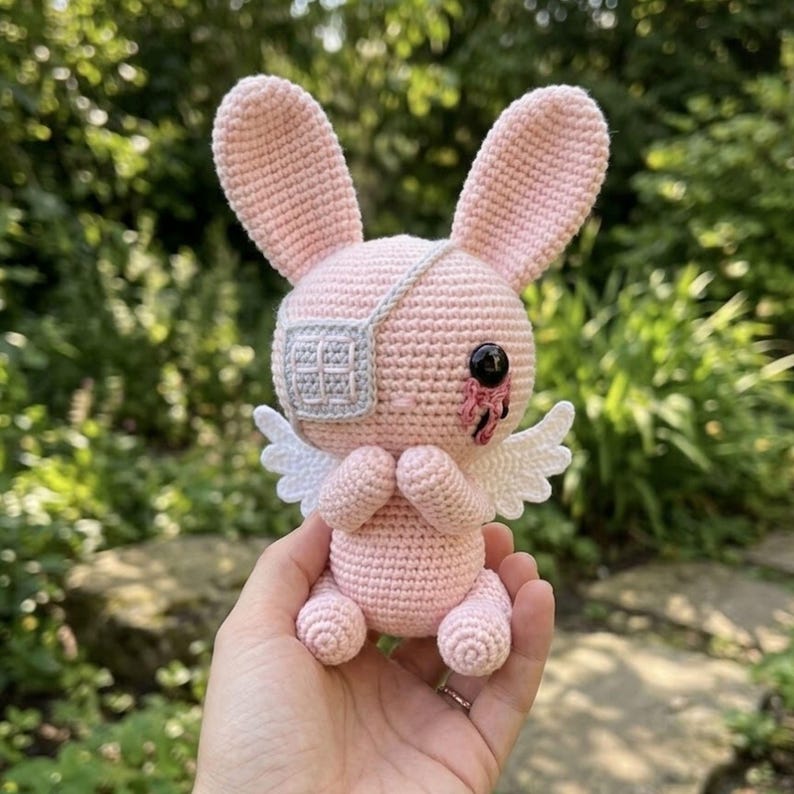 May include: A handmade pink crochet bunny with large ears, a white eye patch, and small white wings. The bunny has a black eye with a red tear and is holding its paws together. A unique, whimsical plush toy.