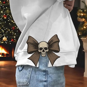 May include: White sweatshirt with a brown bow and skull design. The embroidered bow is large and detailed, with a skull in the center. The sweatshirt is paired with blue jeans.