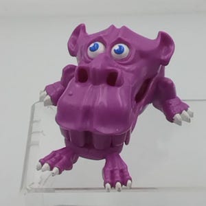 Peut inclure: Figurine de monstre violet avec de grandes dents, des yeux bleus et des griffes blanches. Le jouet a une large bouche et de grandes oreilles. La figurine est posée sur une surface transparente.