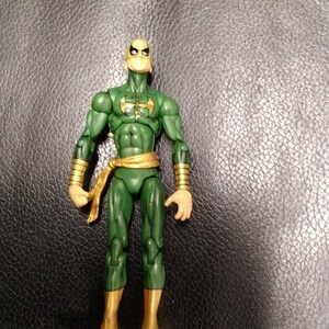Puede incluir: Una figura de acción verde de Iron Fist, un superhéroe de Marvel Comics. La figura tiene una máscara amarilla, detalles dorados en las muñecas y botas, y una faja dorada. La figura está de pie.