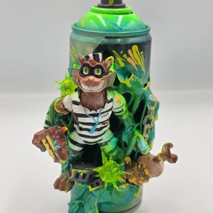 May include: A custom-painted spray paint can featuring a cartoon cat character in a prison uniform. The can is primarily green with yellow accents and a black nozzle. The cat is brown and white, holding a small sign. The overall design has a graffiti art style.