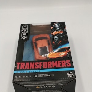 May include: A Transformers Studio Series KSI Widow action figure in its original packaging. The box is black with orange and white text and graphics. The toy car is orange and black. The box also features an image of a robot.