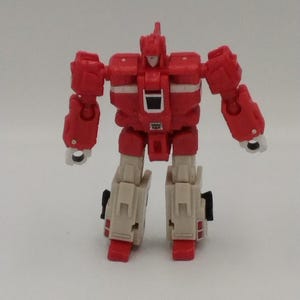 May include: A red and off-white action figure robot with articulated arms and legs. The toy robot has a rectangular torso, a head with a pointed top, and white accents. The figure is standing upright against a plain background.