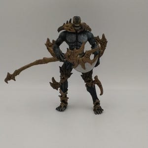 Puede incluir: Figura de acción detallada de una criatura oscura y musculosa con armadura puntiaguda y una expresión amenazante. La figura es principalmente negra y gris, con detalles marrones en la armadura y un arma grande y curvada. La figura está de pie en una pose dinámica.