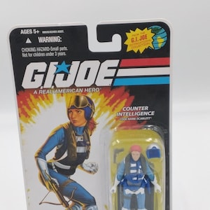 May include: G.I. Joe action figure of Scarlett, a counter-intelligence operative. The figure is in a clear plastic case attached to a black and white card with the G.I. Joe logo and the text "A Real American Hero". The packaging includes a warning about small parts.