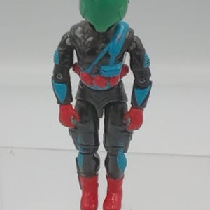 May include: A vintage action figure of a futuristic soldier. The figure is mainly black and grey, with red gloves and boots, and blue accents on the arms and legs. It wears a green helmet and a red belt.
