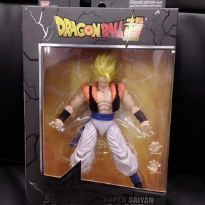 Puede incluir: Figura de acción de Super Saiyan Gogeta de Dragon Ball Super en una caja de exhibición. La figura tiene cabello amarillo, ropa naranja y negra, y pantalones blancos. La caja tiene el logotipo de Dragon Ball Super y "Dragon Stars Series".