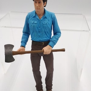 May include: A figurine of a man in a blue button-down shirt, brown pants, and brown shoes, holding a large axe with a wooden handle. The figure has dark hair and is standing in front of a clear, rectangular structure.