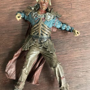 Puede incluir: Figura de acción detallada de un personaje parecido a un zombi. La figura lleva un casco adornado con calaveras, ropa hecha jirones y una capa marrón. La figura tiene una placa torácica esquelética y posa con los brazos extendidos.