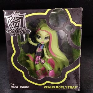 Peut inclure: Figurine en vinyle de Venus McFlytrap dans une boîte. La poupée a la peau verte, de longs cheveux verts avec des mèches roses et porte une tenue colorée. La boîte affiche le logo Monster High et le texte "8+ VINYL FIGURE".
