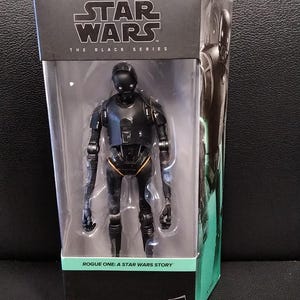 Puede incluir: Figura de acción negra K-2SO de la serie Star Wars Black, empaquetada en una caja de plástico transparente. La caja presenta el logotipo de Star Wars y el texto "ROGUE ONE: A STAR WARS STORY". La figura mide unos 15 cm de altura.