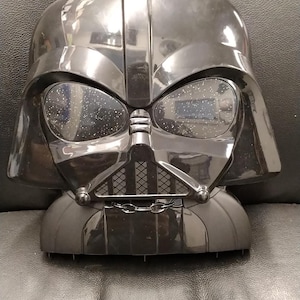 May include: A black Darth Vader helmet-shaped clock. The clock has a glossy finish and features the iconic helmet design with eye lenses and a detailed mouth grill. The clock is a collectible item from the Star Wars franchise.