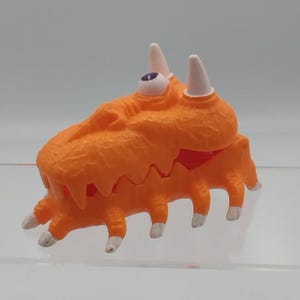 Peut inclure: Une créature jouet orange avec un seul œil, des cornes blanches et plusieurs pattes. Le jouet a une surface texturée et un intérieur rouge visible dans la zone de la bouche. Les pattes ont des pointes blanches.