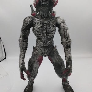 Puede incluir: Figura detallada de una monstruosa criatura alienígena. La figura es principalmente gris con acentos rojos, con un diseño esquelético detallado y una expresión amenazante. La criatura está de pie con las piernas separadas, los brazos a los lados y una cabeza grande.
