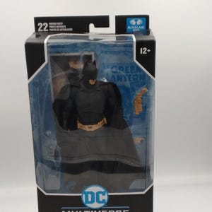 Puede incluir: Figura de acción de Batman con un traje negro y gris, empaquetada en una caja negra y azul. La caja presenta el logotipo de DC Multiverse y el texto "Batman". La figura tiene 22 puntos de articulación y es para mayores de 12 años.
