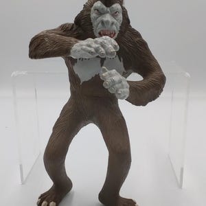 Puede incluir: Figura de monstruo marrón y blanco con expresión amenazante. La figura tiene cuerpo peludo, cara gris claro y manos blancas. El monstruo está de pie con los brazos levantados y la boca abierta, revelando encías y dientes rojos.