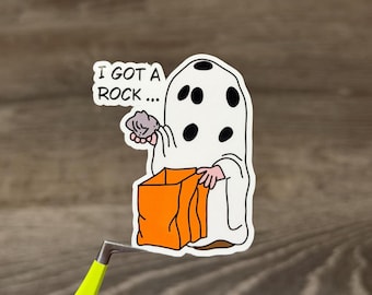 May include: A die-cut vinyl sticker featuring a cartoon character dressed in a white ghost costume with black polka dots, holding a gray rock and an orange trick-or-treat bag. The text bubble above the character reads, I GOT A ROCK...