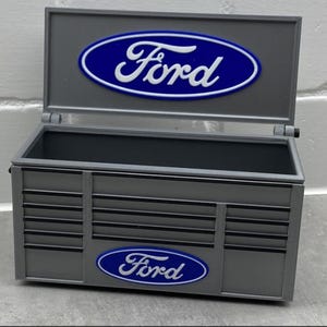 Può includere: Una cassetta degli attrezzi grigia in miniatura con il logo Ford bianco su un ovale blu. La cassetta degli attrezzi ha un coperchio incernierato e un design a cassetto sul davanti. Il logo Ford è anche sul davanti.