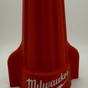Könnte beinhalten: Ein roter Elektrokabelverbinder mit geripptem Design. Das Wort "Milwaukee" ist in Weiß mit einem Blitz aufgedruckt. Der Verbinder hat einen ausgestellten Sockel und eine konische Oberseite.