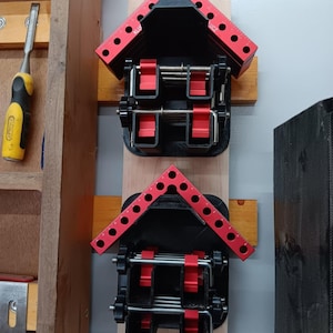May include: Two red and black corner clamps mounted on a wooden wall. The clamps are used to hold pieces of wood together at a 90-degree angle.