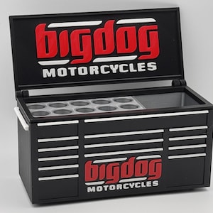 Può includere: Una cassetta degli attrezzi in metallo nero con coperchio incernierato che riporta la scritta rossa e bianca Big Dog Motorcycles. Il coperchio aperto rivela un vassoio interno grigio con dodici scomparti circolari. Il frontale presenta vari cassetti.