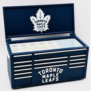 Può includere: Una scatola blu navy con il logo e il testo dei Toronto Maple Leafs. La scatola ha un coperchio incernierato e una maniglia bianca. L'interno ha una sezione con scomparti circolari. Il logo è una foglia d'acero bianca.