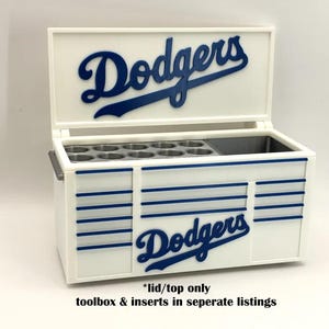 Può includere: Contenitore termico bianco e blu a tema Dodgers, con il logo della squadra sul coperchio e sulla parte anteriore. Il contenitore termico ha una forma rettangolare con strisce blu e una maniglia. Il coperchio è aperto, rivelando un vano con inserti circolari.