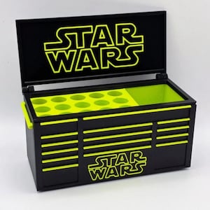 Può includere: Cassetta degli attrezzi nera con dettagli verde neon e il logo di Star Wars. La cassetta ha un coperchio incernierato e un vano interno verde neon con fori circolari. Il logo di Star Wars è anche sulla parte anteriore.