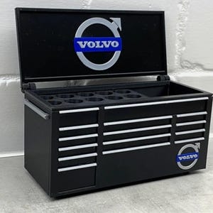 Può includere: Cassetta degli attrezzi nera in miniatura con dettagli bianchi e logo Volvo. La cassetta ha più cassetti e un vano superiore. Il logo Volvo è visualizzato sulla parte superiore e laterale. La cassetta è aperta, rivelando l'interno.