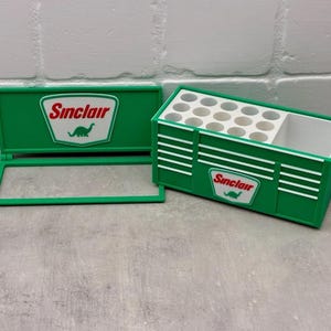 Può includere: Una scatola portaoggetti verde con coperchio incernierato e scomparto separato. La scatola presenta il logo Sinclair con un dinosauro in un emblema bianco e rosso. La scatola ha strisce orizzontali bianche e una serie di fori circolari.