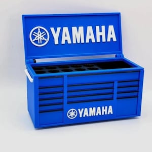 Può includere: Una cassetta degli attrezzi Yamaha blu con coperchio incernierato e più cassetti. Il logo e il testo Yamaha sono bianchi sul coperchio e sulla parte anteriore. La cassetta degli attrezzi ha una maniglia bianca e un interno nero.