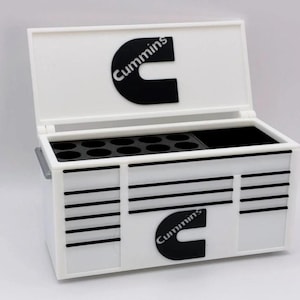 May include: White storage box with a hinged lid, featuring the "Cummins" logo in black. The box has multiple compartments and drawers, with the logo repeated on the front. The box is designed for organization and storage.