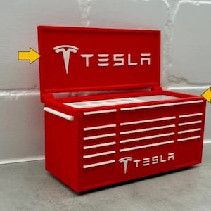 Può includere: Una cassetta degli attrezzi rossa con il logo e il testo Tesla in bianco. La cassetta degli attrezzi ha un coperchio incernierato e più cassetti. Il logo Tesla è anche sulla parte anteriore. La cassetta degli attrezzi è su una superficie grigia.