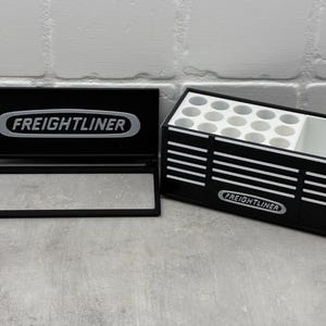 Può includere: Organizzatore nero con il logo Freightliner. Ha una forma rettangolare con più scomparti per riporre oggetti. Il coperchio è aperto, rivelando l'interno. L'organizzatore è su una superficie grigia.