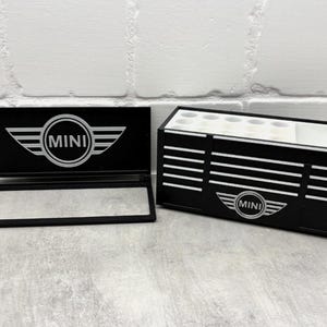Può includere: Organizzatore nero e bianco con marchio MINI e una scatola abbinata con parte superiore aperta. L'organizzatore ha un coperchio incernierato con il logo MINI. La scatola ha più scomparti e il logo MINI. Gli articoli sono su una superficie grigio chiaro.