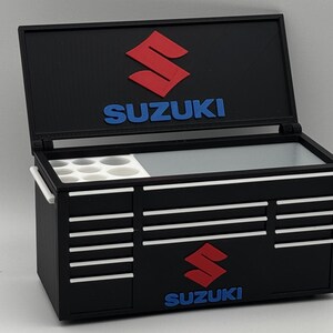 Può includere: Una cassetta degli attrezzi in miniatura nera con coperchio incernierato che presenta il logo Suzuki rosso e testo blu. L'interno include un vassoio bianco con fori circolari. L'esterno mostra il logo e il testo Suzuki, con dettagli bianchi sulle maniglie.