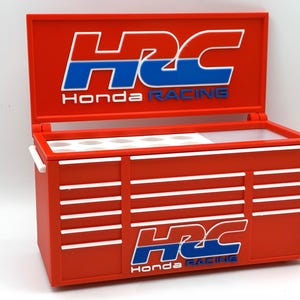 Può includere: Una cassetta degli attrezzi in miniatura rosso brillante con coperchio incernierato che presenta il logo HRC Honda Racing in blu e bianco. La cassetta ha diversi frontali di cassetti orizzontali con finiture bianche e un vassoio interno bianco.